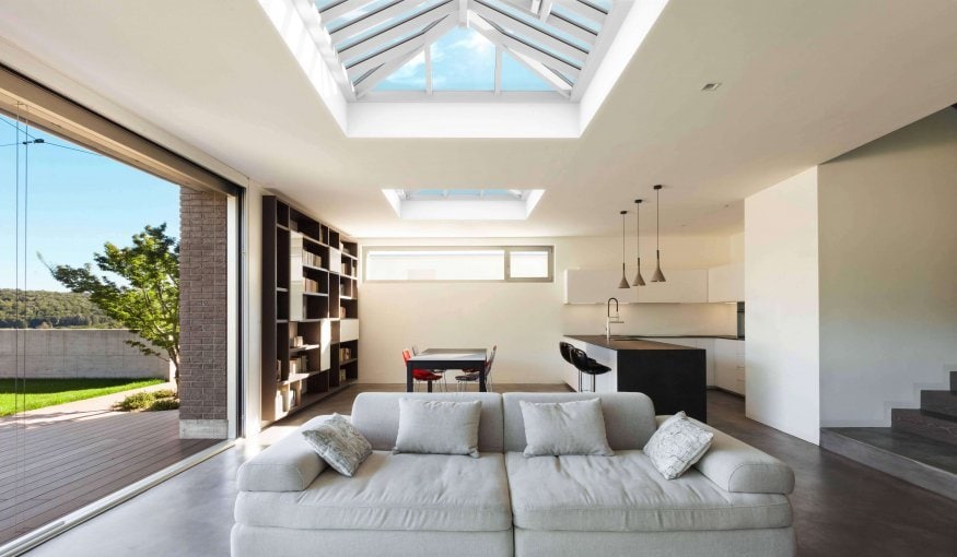 Modern open-plan living room and kitchen with skylights, large sofa, and sliding glass doors leading to a patio and greenery.