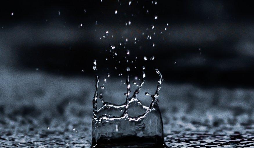 A water droplet splashes on a dark surface, creating ripples and scattered droplets in mid-air.